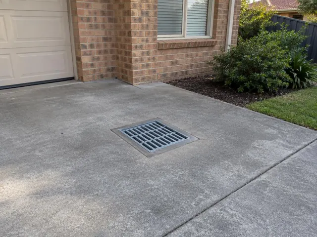 Dry concrete driveway with a square metal drain grate beside a brick house in Kings Park