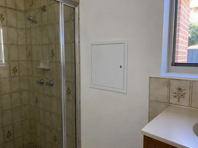 Bathroom wall with a small access panel beside tiled shower and window in Lalor Park